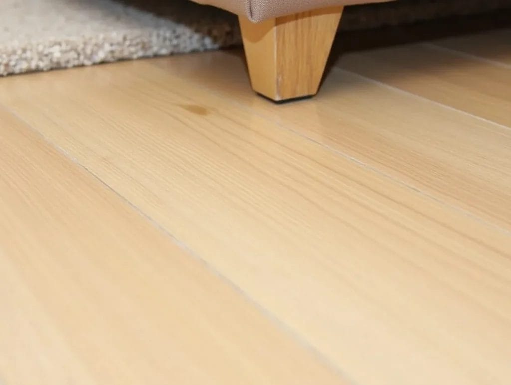 A close-up of a light oak floor with slight wear, taken from a skewed angle, showing part of a sofa leg and a carpet
